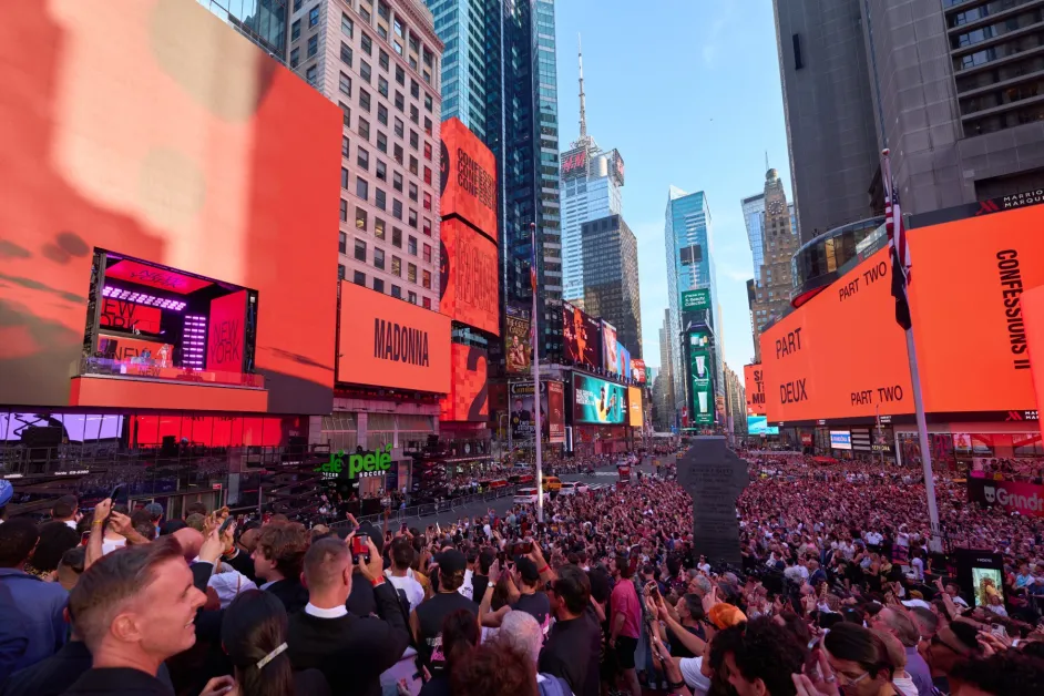 A massive crowd fills Times Square as billboards spelling MADONNA, CONFESSIONS II, PART DEUX and PART TWO glow red above the stage.